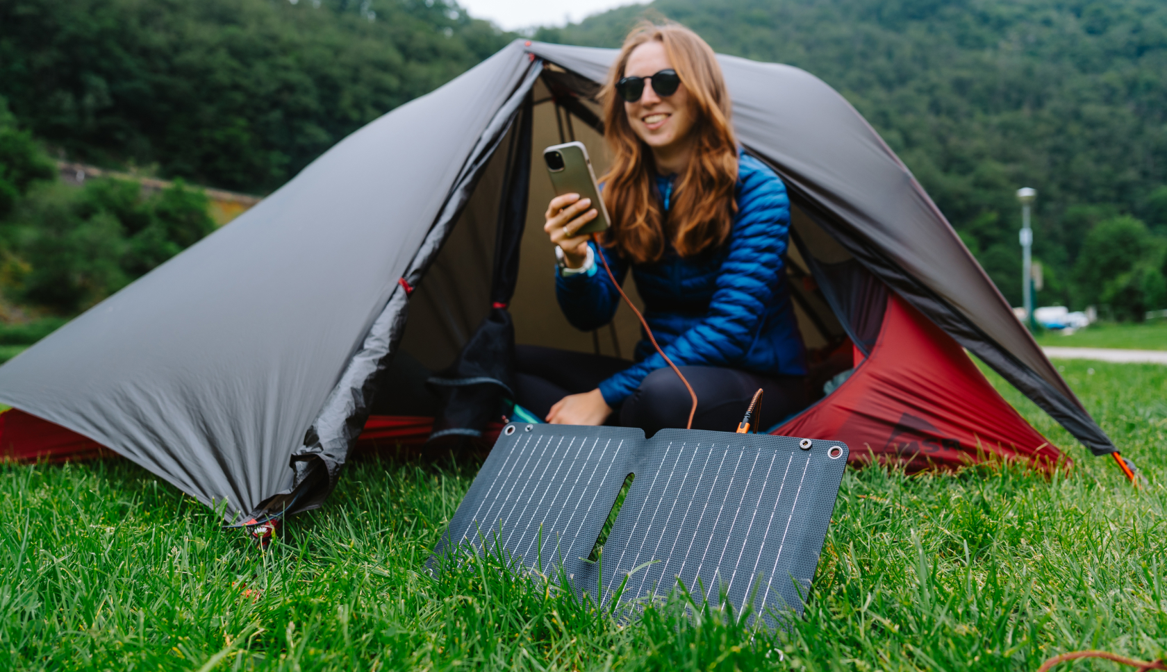 Een vrouw zit voor haar tent en laadt haar telefoon op met een draagbaar zonnepaneel.