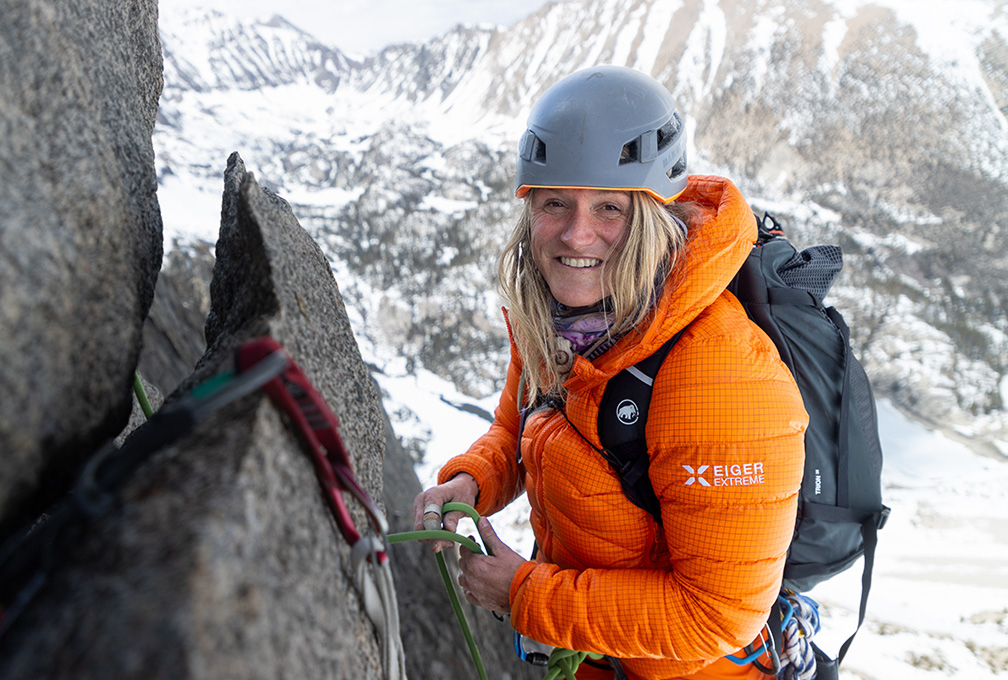 vrouw in klimuitrusting hangt aan een rots met besneeuwde bergen in de verte