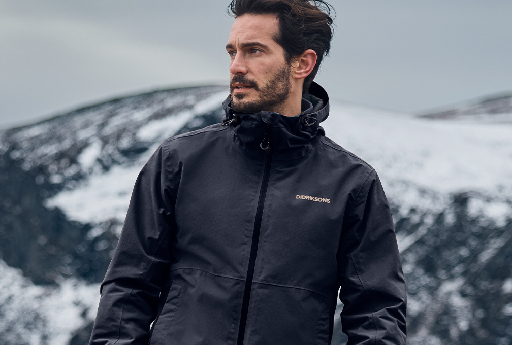Man draagt een zwarte Didriksons-regenjas in een besneeuwd berglandschap.