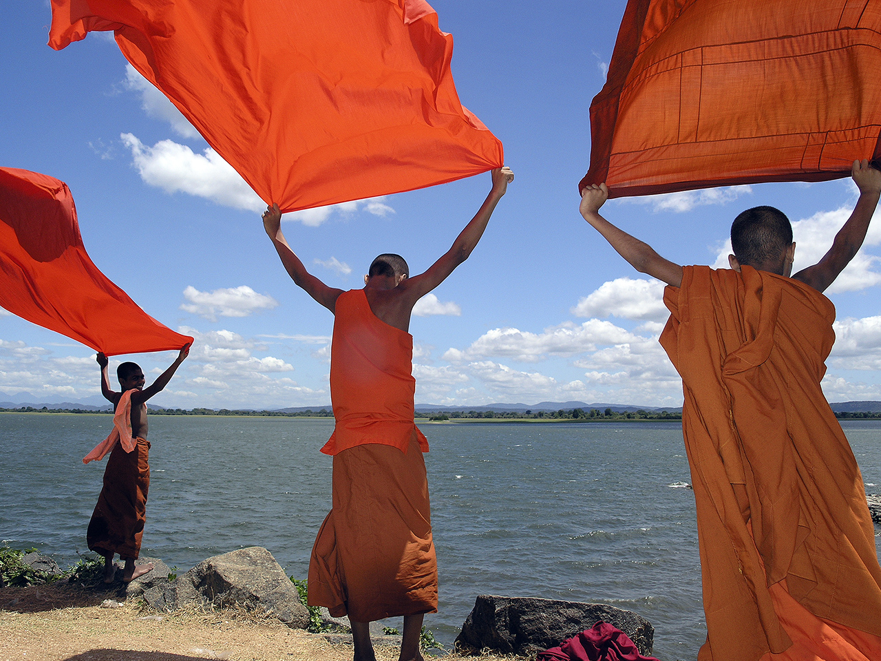 Sawadee - three Buddhist monks standing on the shore of a lake holding up banners
